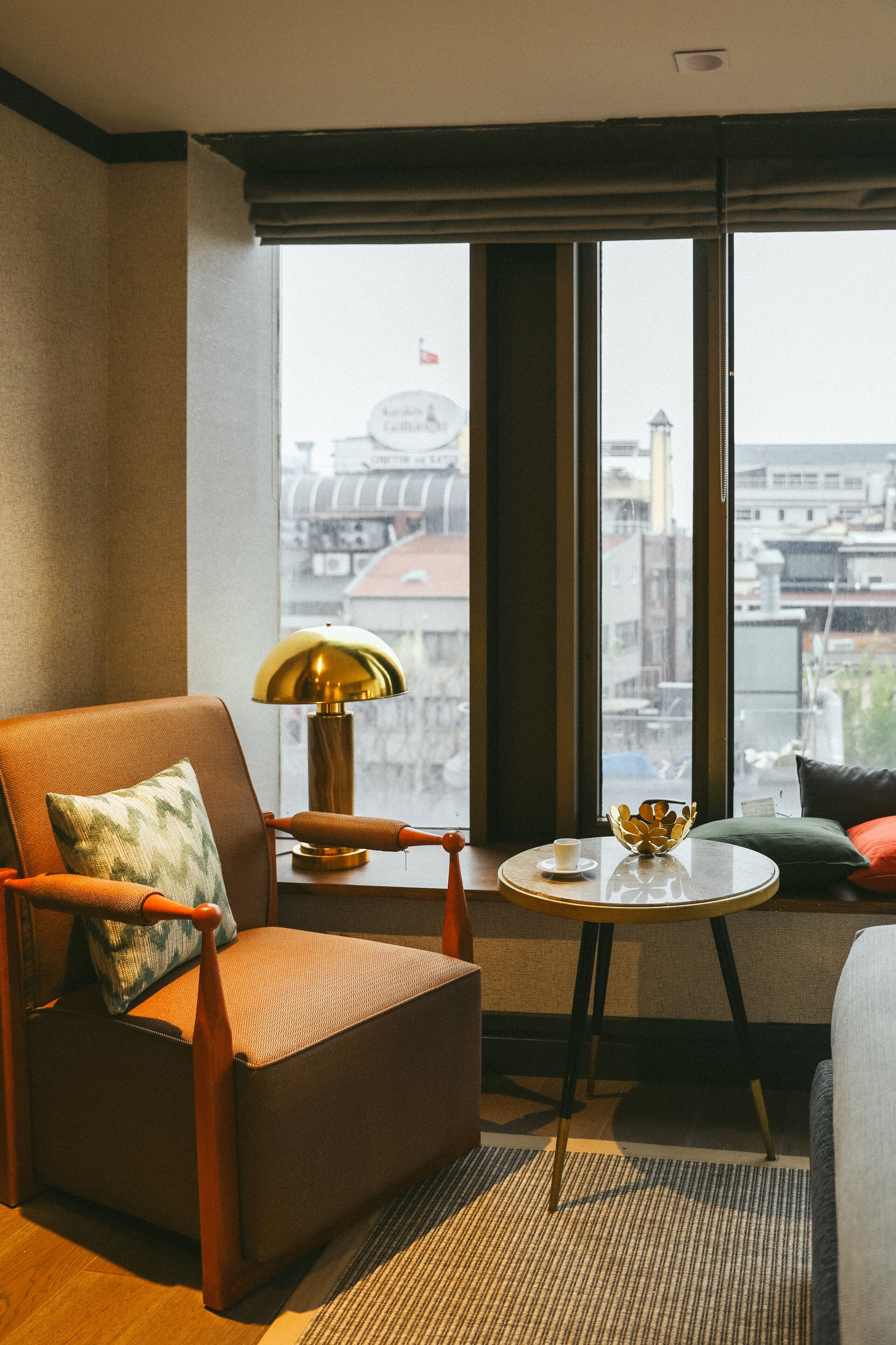 Habitación de hotel con un ventanal grande, una silla, una mesa con lámpara y vista a la ciudad.