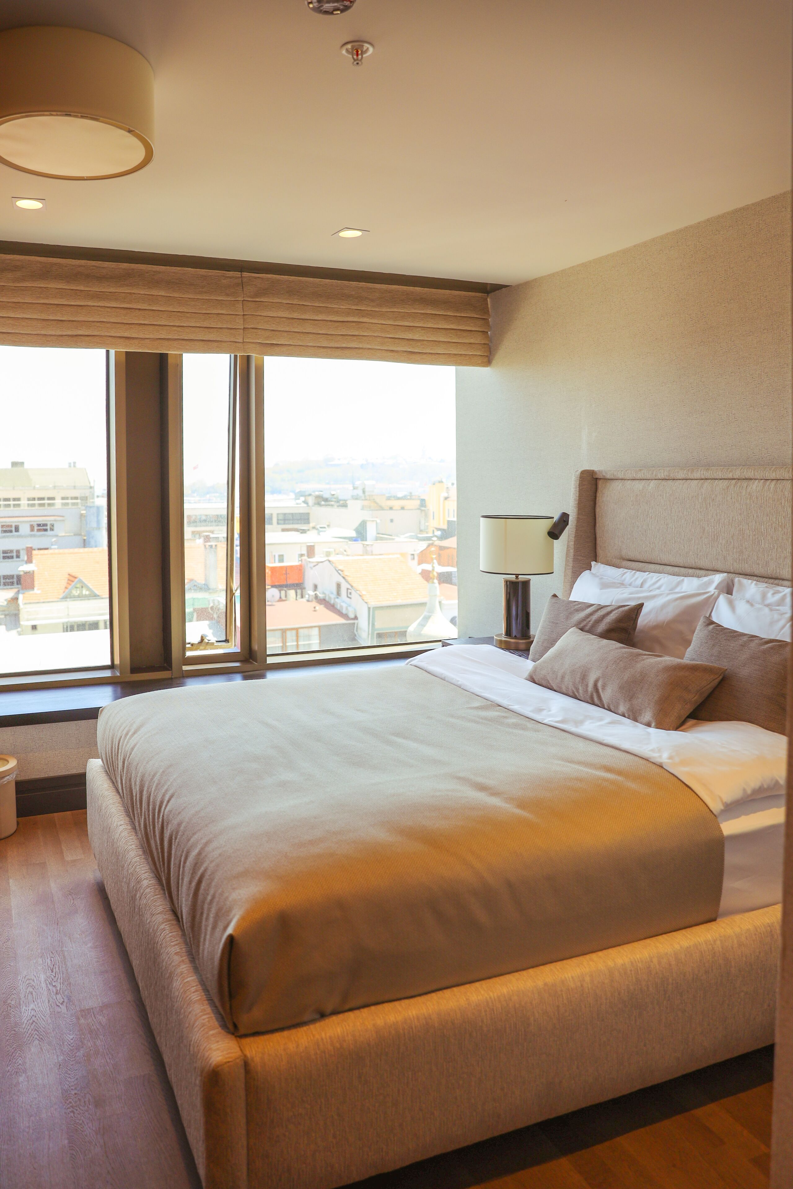 Una cama bien hecha con cabecera, una lámpara de noche y vistas a edificios a través de amplios ventanales.