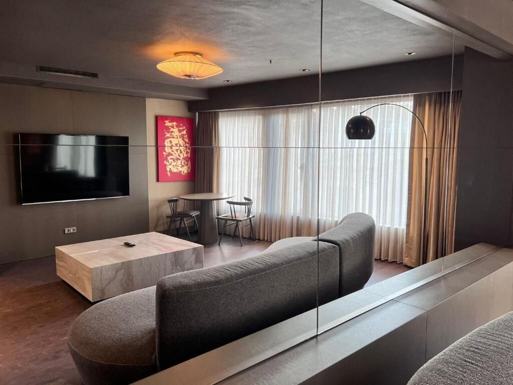 A modern living room with a flat-screen TV, a wooden coffee table, a grey sofa, and a dining area with a table and chairs.