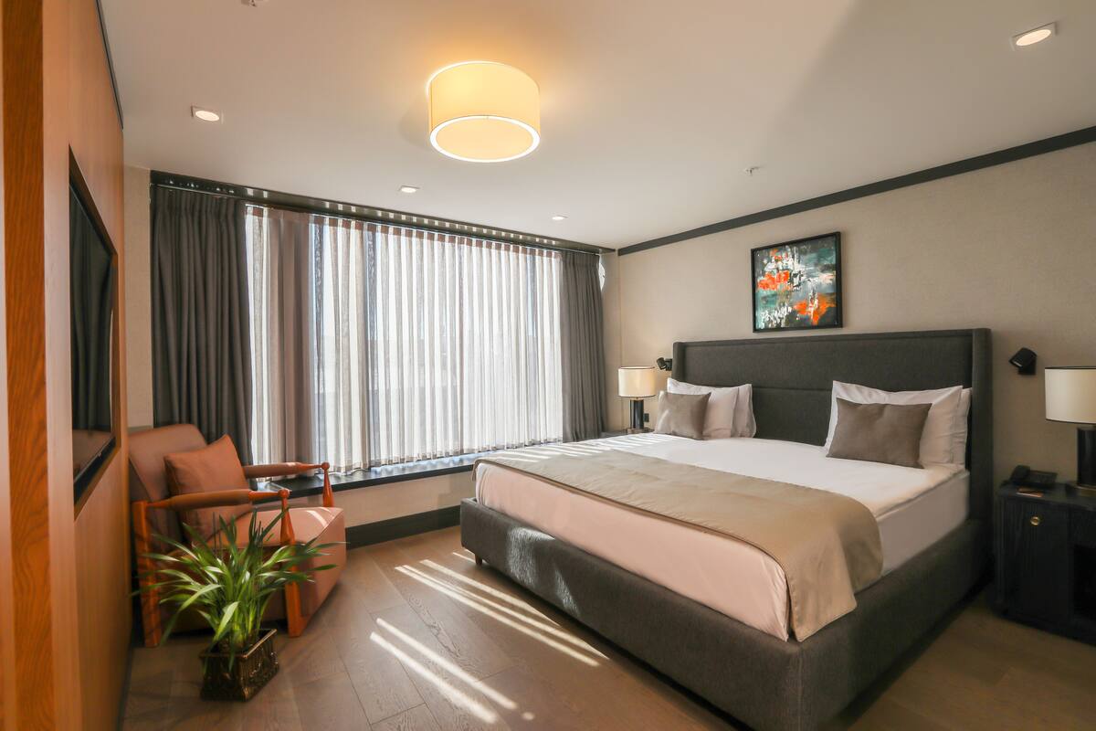 A modern hotel room with a large bed, a seating area with a chair and a table, a potted plant, and a painting on the wall.