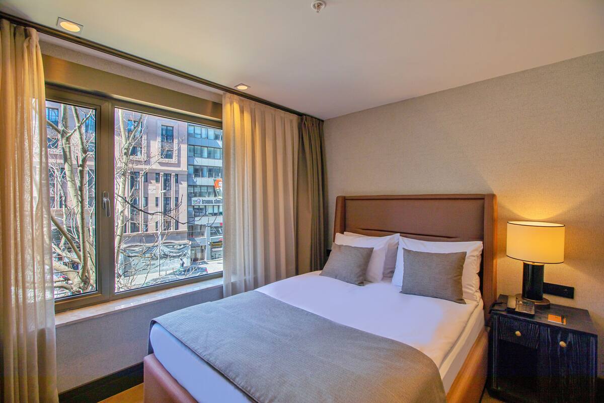 A hotel room with a large bed, a nightstand with a lamp, and a window with curtains.