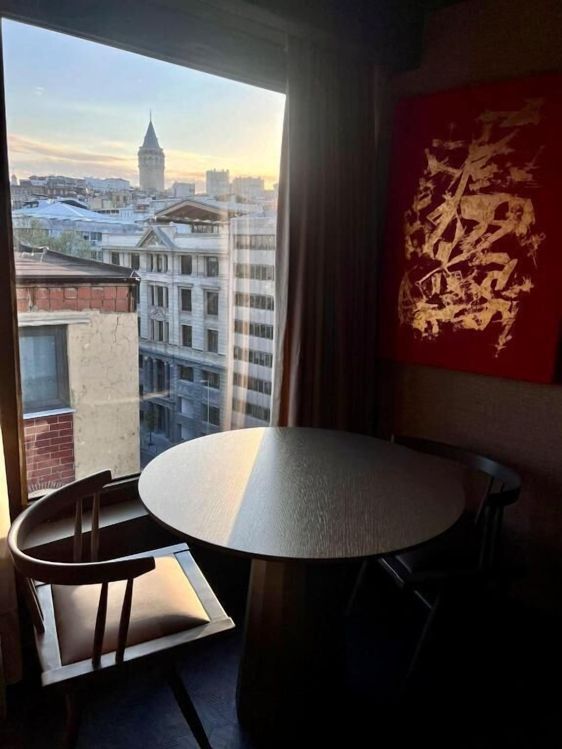 Une pièce avec une table ronde et deux chaises, une grande fenêtre donnant sur un paysage urbain comprenant une tour, et une œuvre d’art abstraite rouge accrochée au mur.