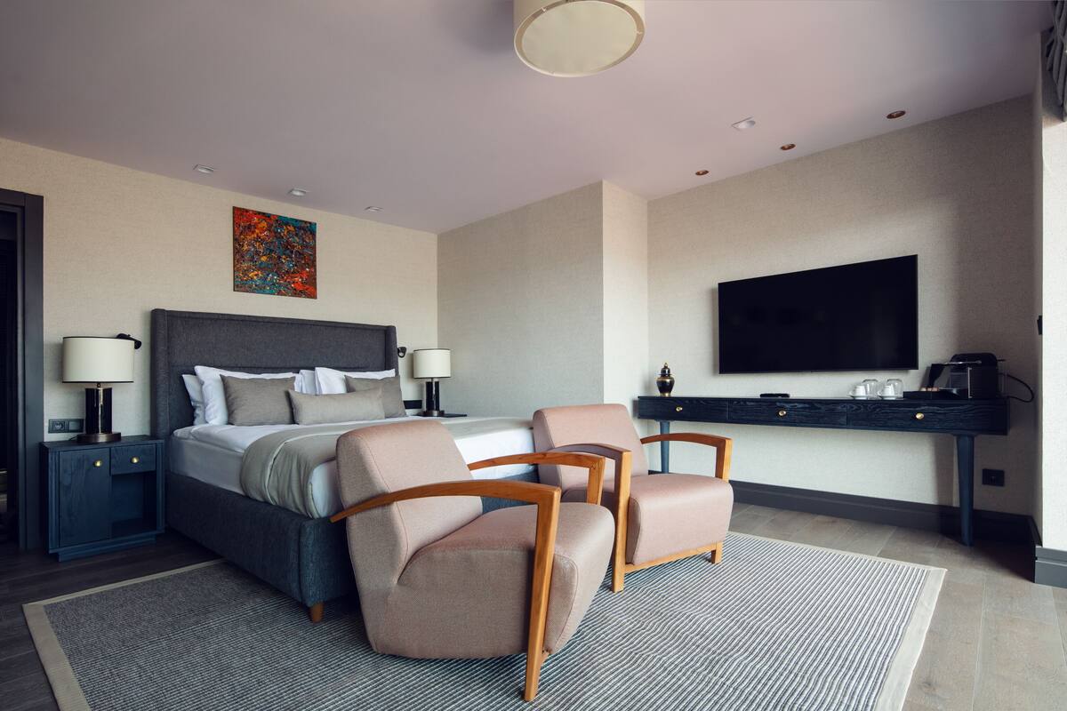 A modern hotel room with a large bed, two armchairs, a flat-screen TV, and a coffee machine on a dark console.