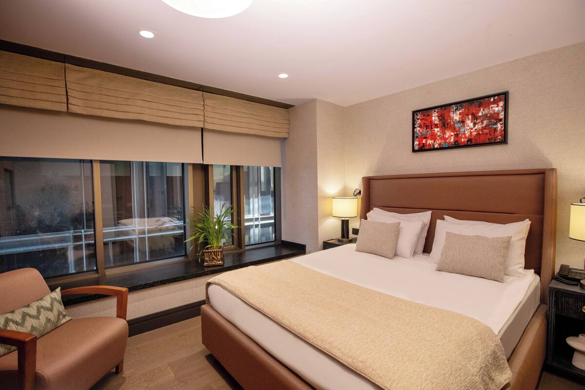 A hotel room with a large bed, a sofa, a window with blinds, a bedside table with a lamp, and a wall-mounted painting.