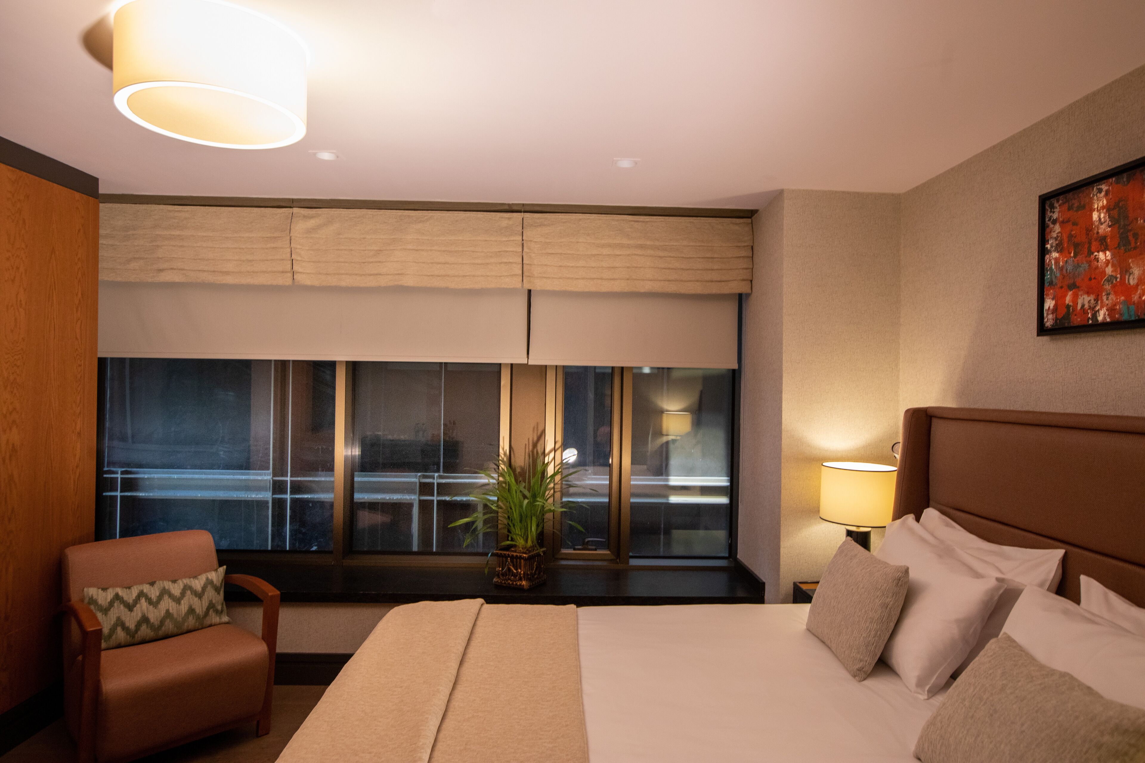 Une chambre d’hôtel avec un grand lit, une chaise, une fenêtre avec des stores, une plante et un tableau au mur.