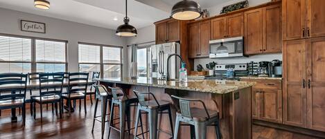 A modern kitchen with wooden cabinets, a central island with bar stools, stainless steel appliances, and a view through large windows.