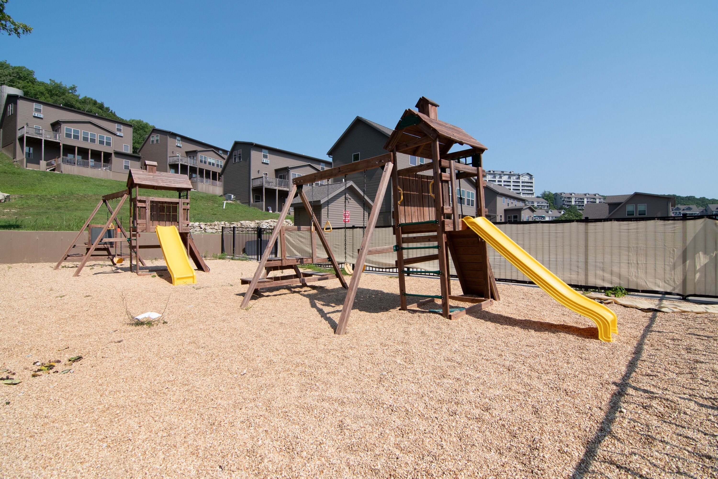 A playground with wooden structures and yellow slides, set against a backdrop of residential buildings.
