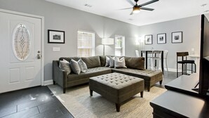 Un salon moderne avec un canapé d’angle, un pouf, un ventilateur de plafond et un tapis de course.