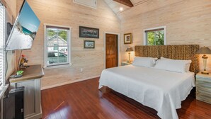 Une chambre avec un plafond en bois, un grand lit, un téléviseur fixé au mur et une fenêtre avec des stores.