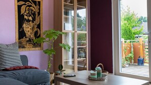 Een kamer met een paarse wand, een groot wandtapijt, een houten kast, een bank en een raam met uitzicht op groen.