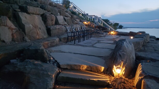 A stone pathway leading to a seating area with chairs and a fire pit, overlooking a body of water.