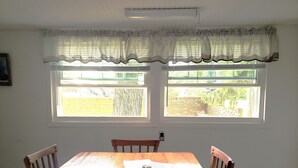 A dining area with a wooden table and chairs, two windows with a view of a brick wall and greenery, and a white ceiling with a light fixture.