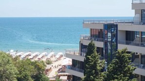 A beachfront hotel with multiple balconies overlooking the sea, a pool area with umbrellas, and a clear view of the ocean.
