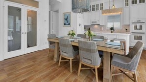 A modern kitchen with white cabinetry, a wooden dining table, wicker chairs, and a blue chandelier. - NEAPOLITAN SKY - Key West Style Home in Olde Naples (Naples)