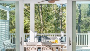 A covered patio area with a table set for two, outdoor furniture, and a view of a wooded area. - Modern coastal retreat steps from Center Street and the beach (Folly Beach)