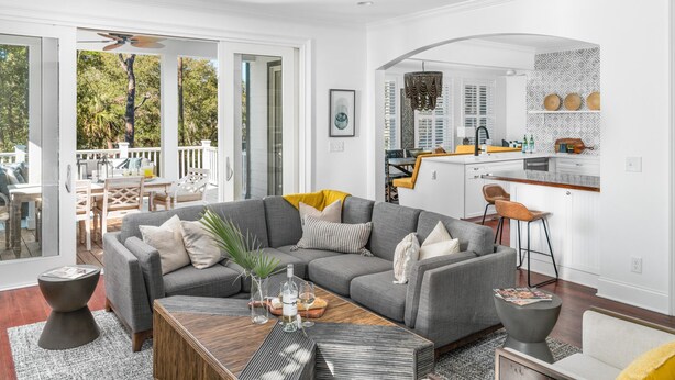 A modern living room with a grey sectional sofa, a wooden coffee table, and a kitchen area with white cabinets and a bar stool. - Modern coastal retreat steps from Center Street and the beach (Folly Beach)