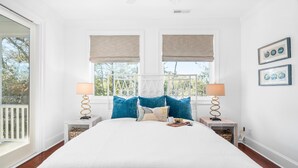 A bedroom with a large bed, two bedside tables, a window with blinds, and a view of the outdoors. - Modern coastal retreat steps from Center Street and the beach (Folly Beach)