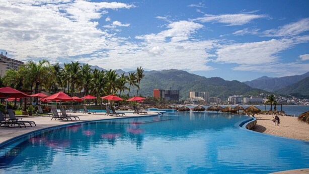 A pool area with red umbrellas, lounge chairs, and a view of mountains and buildings in the background. - Beachfront 1 br. condo with private hot tub at Gran Venetian Puerto Vallarta (Puerto Vallarta)