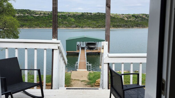 A wooden deck with two black chairs, a dock leading to a green boat house, and a lake view. - Wet Feet Retreat! Lake front cabin in quiet cove. (Caddo)