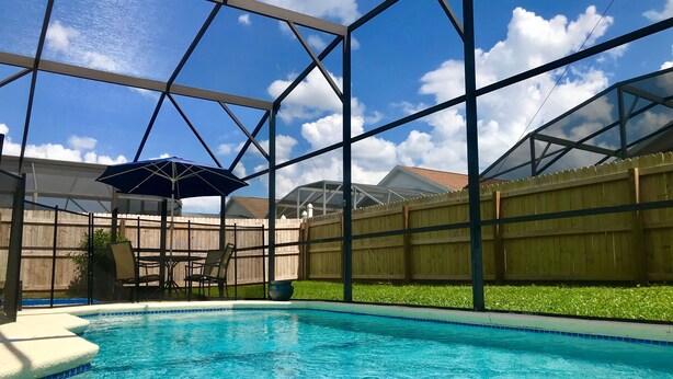 Ein Swimmingpool mit blauem Himmel und Wolken im Hintergrund, umgeben von einem Holzzaun und einer überdachten Terrasse.