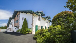 Il s’agit d’un bâtiment à deux étages, doté d’un toit en pente, de murs extérieurs blancs et de volets verts. Un grand conifère se trouve devant le bâtiment.