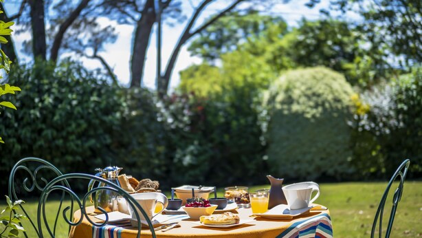 Une table de jardin, dressée pour le petit-déjeuner, entourée de verdure et d’arbres.