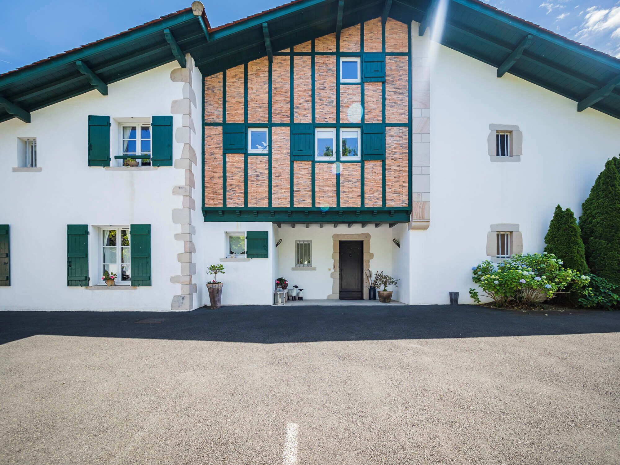 Edificio de dos pisos con fachada blanca y contraventanas verdes, que presenta una sección central de ladrillo y una disposición simétrica de ventanas.
