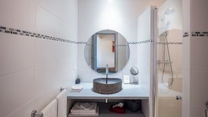Une salle de bain moderne avec des murs recouverts de carreaux blancs, un miroir rond, un lavabo rond, une douche avec une porte en verre et un petit tabouret.