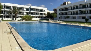 A rectangular swimming pool with a tiled deck, surrounded by white buildings with balconies and a clear blue sky. - Tossan House in Vilamoura Downtown (Quarteira)