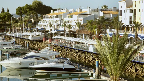 A marina with several boats docked, a stone wall, and buildings with balconies and outdoor seating. - Tossan House in Vilamoura Downtown (Quarteira)