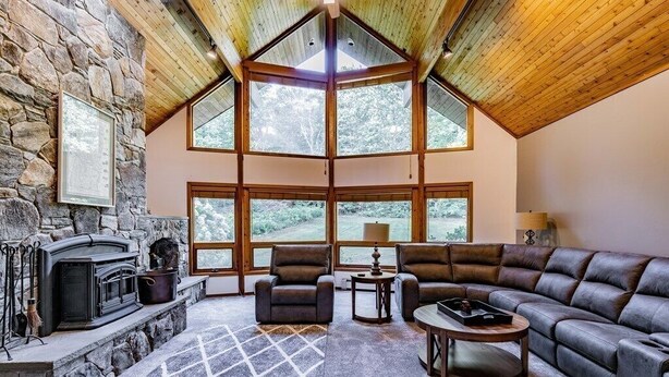 A cozy living room with a stone fireplace, leather sofas, and large windows offering a view of the outdoors. - Wildlife Retreat in the Forest (Becket)