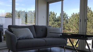 Ett modernt vardagsrum med en grå soffa, ett matbord och stora fönster som ger utsikt över träd och en klar himmel.