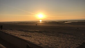 En strand med en solnedgång i bakgrunden, människor som promenerar längs stranden och ett lugnt hav.
