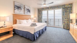 Ein Hotelzimmer mit einem großen Bett, einem Deckenventilator, Blick auf den Ozean und einem Balkon mit gemusterten Vorhängen.