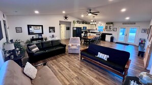 A spacious living room with a black leather sofa, a beige sofa, and a dining area with a wooden table and chairs. There is a kitchen in the background with white cabinets and a black refrigerator.
