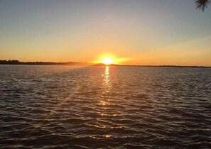 Un coucher de soleil sur une étendue d’eau calme, avec une côte visible au loin.