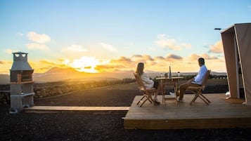 Un couple est assis à une table en bois, face à un paysage montagneux illuminé par le coucher du soleil.