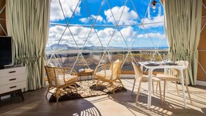 A room with a large window offering a mountain view, wicker furniture, a small table, and a TV on a cabinet.