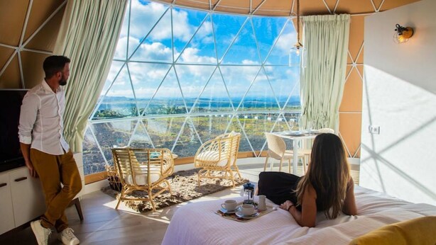 A bedroom with a large window, a bed, a table with chairs, and a TV.