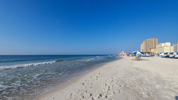A beach with footprints leading to the water, a clear sky, and buildings in the background. - Harbour Place 204 - white sands, and beach view! (Orange Beach)