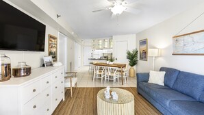 A modern living room with a blue sofa, a coffee table, a TV, and a dining area with a table and chairs. - Harbour Place 204 - white sands, and beach view! (Orange Beach)