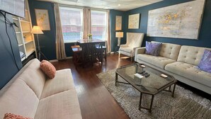 A living room with a sofa, a coffee table, a chair, and a dining table. There are two windows with curtains, a lamp, and a painting on the wall. - On Main Street - Apartment C  - Berlin, MD (Berlin)