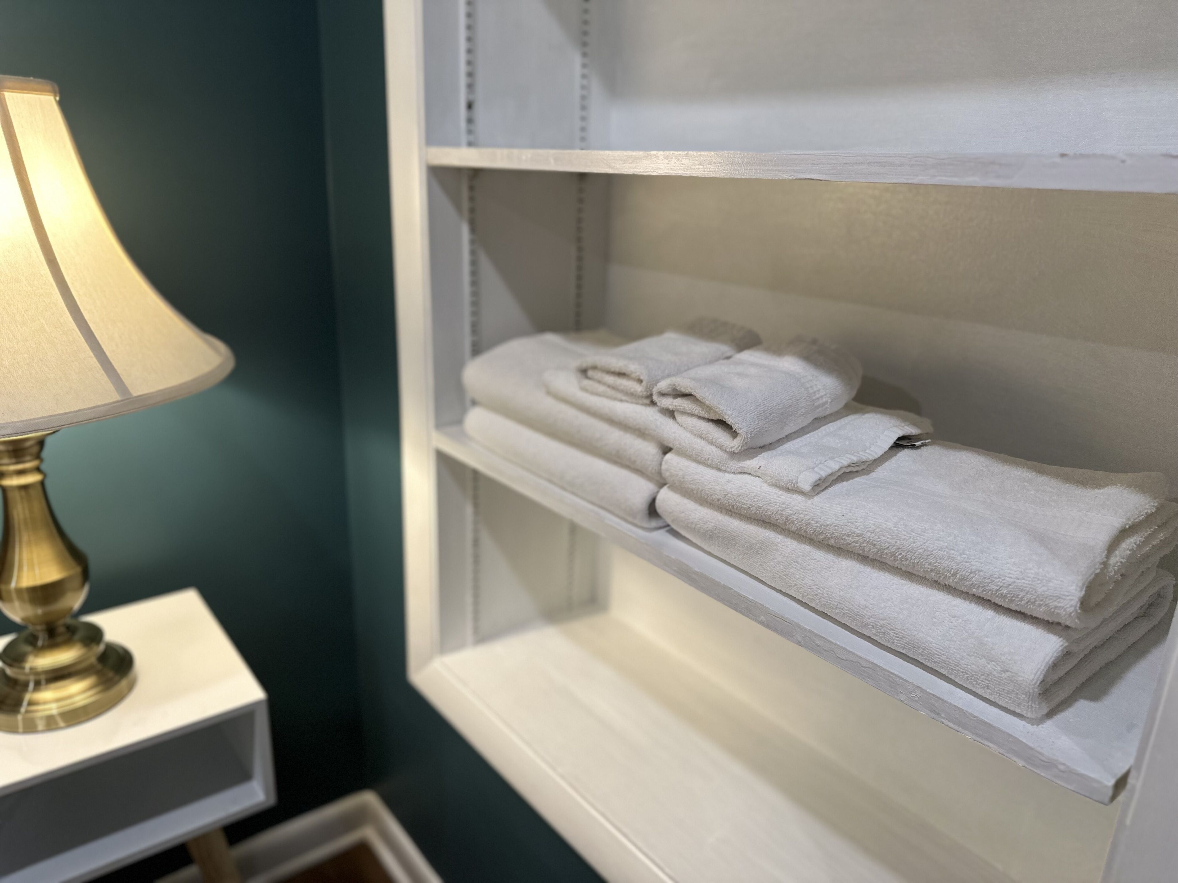Un baño con un estante blanco que sostiene toallas dobladas y una lámpara de latón sobre una mesita de noche blanca.