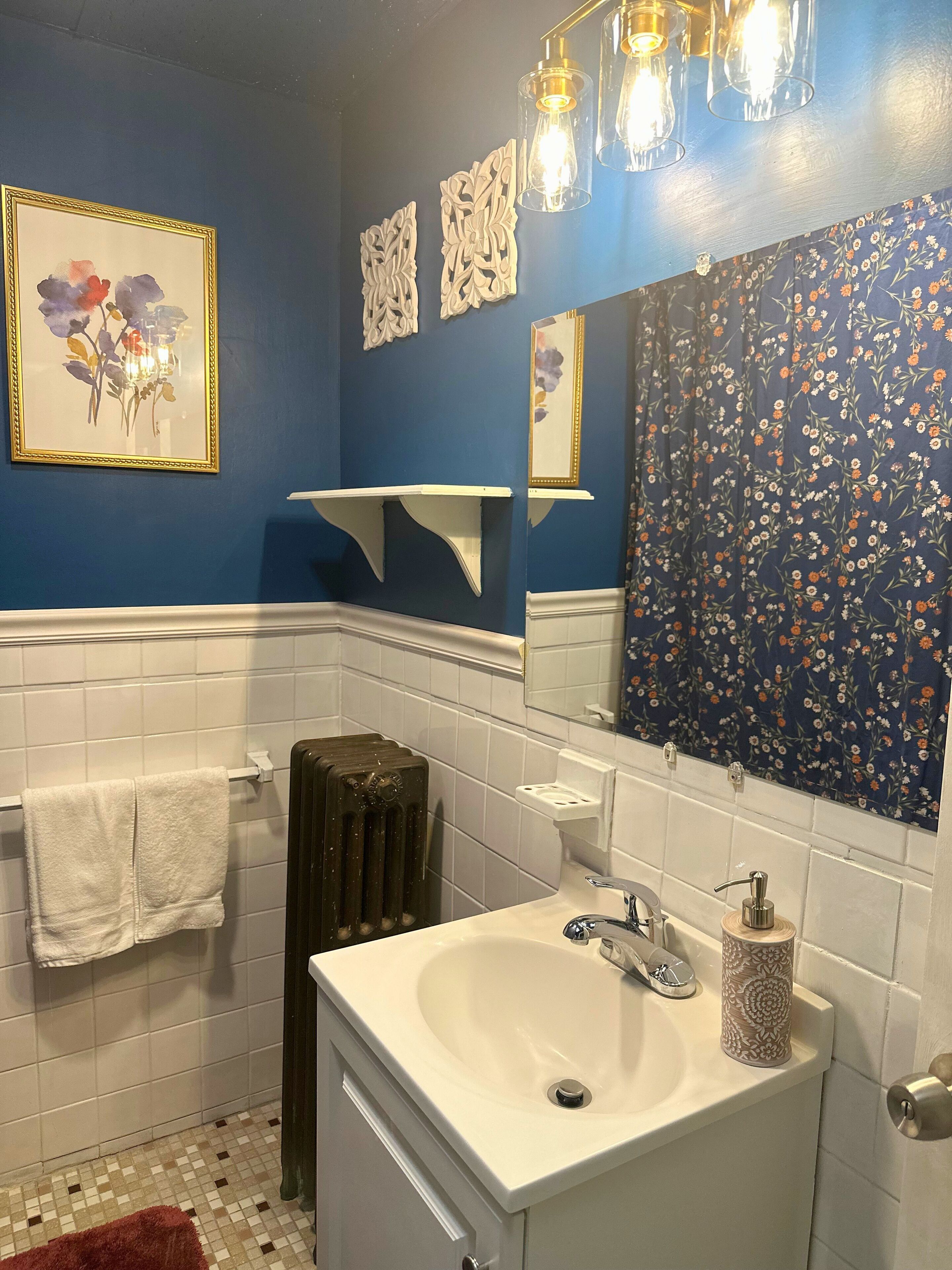 Une salle de bain avec un lavabo blanc, un miroir agrémenté d’un rideau à motifs floraux et un radiateur.