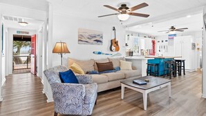 A spacious living area with a sofa, coffee table, and patterned armchair. A guitar hangs on the wall, and there are ceiling fans. - Dolphin Cove • Dazzling Waterfront Cottage w/ Dock (Ocean Isle Beach)
