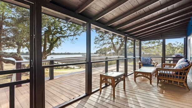 A covered outdoor deck with wooden flooring, glass railings, wicker furniture, and a view of trees and a distant landscape. - Dolphin Cove • Dazzling Waterfront Cottage w/ Dock (Ocean Isle Beach)