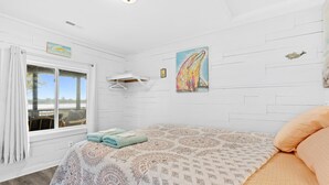 A bedroom with a bed, a window with curtains, a wall-mounted drying rack, and a painting of a whale. - Dolphin Cove • Dazzling Waterfront Cottage w/ Dock (Ocean Isle Beach)
