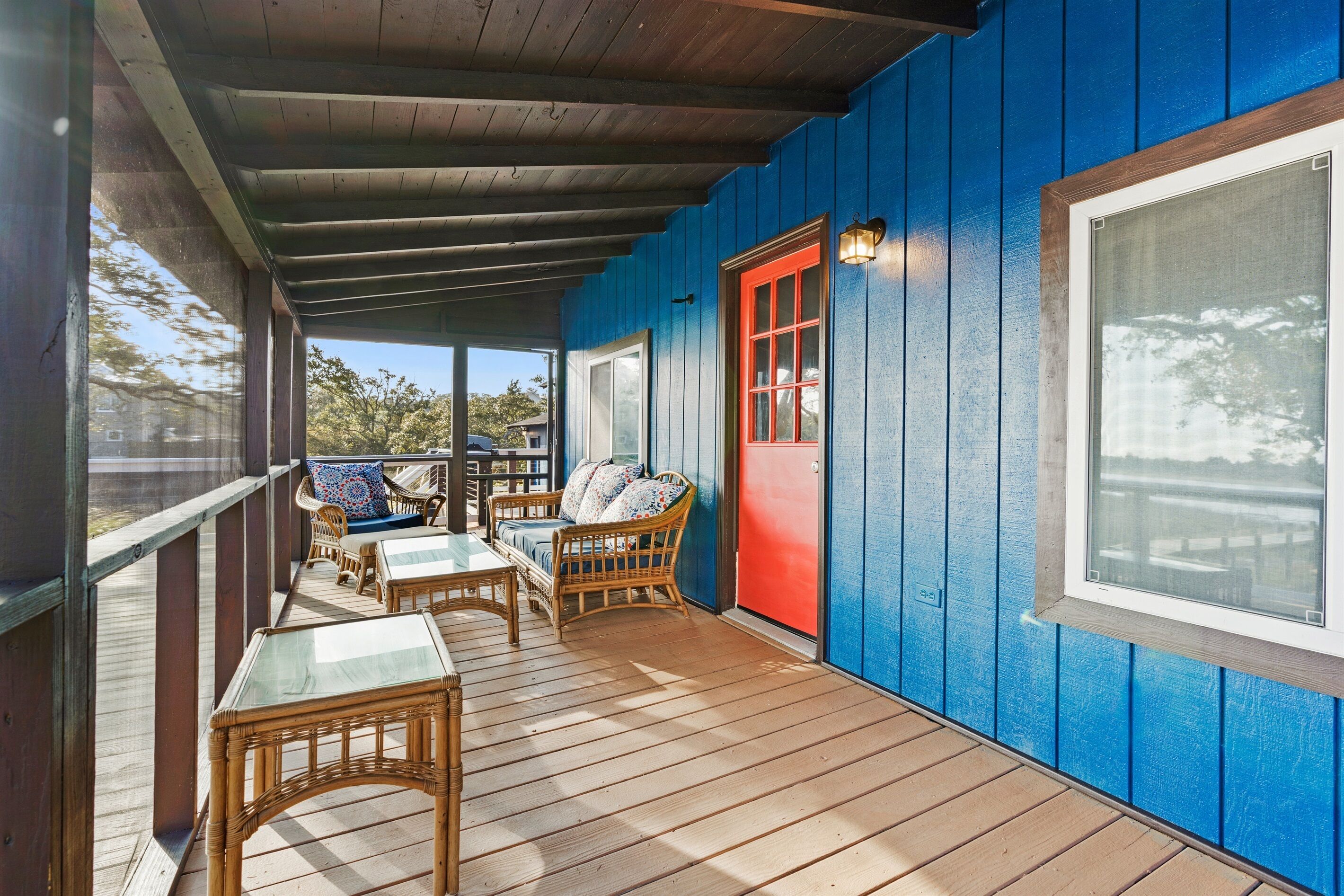 A covered porch with wooden flooring, wicker furniture, and a red door.