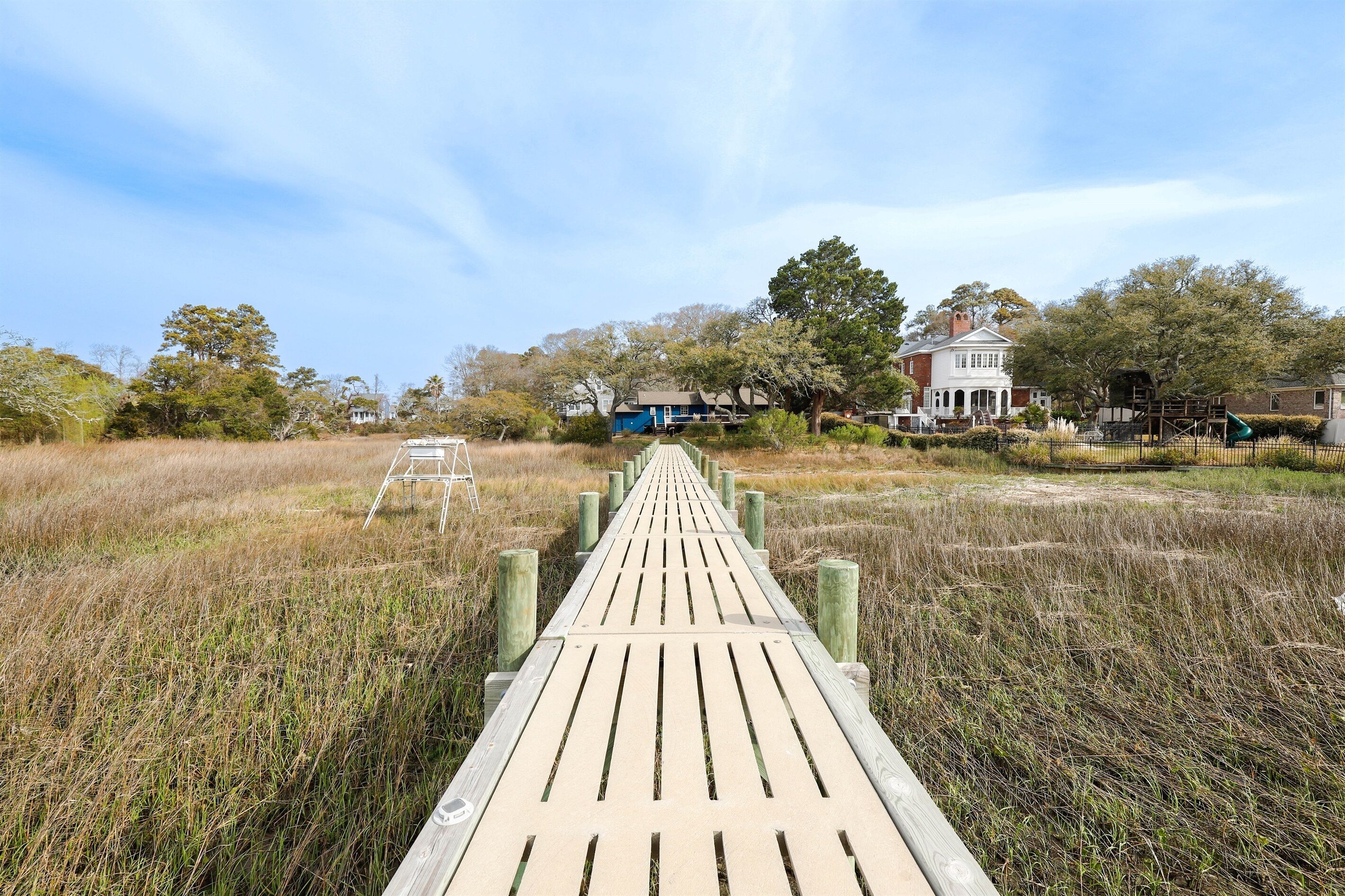 A wooden boardwalk leading to a white house with a blue door, surrounded by tall grass and trees.