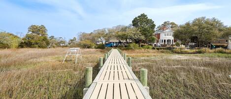 A wooden boardwalk leading to a white house with a blue door, surrounded by tall grass and trees.